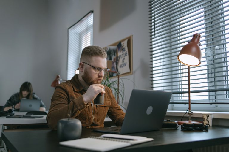 Junger Mann hält Kaffee in der Hand und schaut ernst auf Laptop