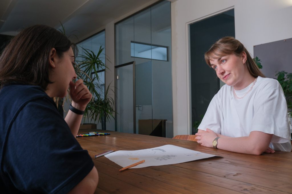 Zwei Frauen sitzen beim Startup-Coaching an Holztisch, zwischen ihnen eine Zeichnung